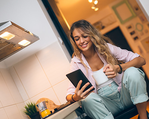 smiling woman drinking coffee in kitchen while reading on her phone
