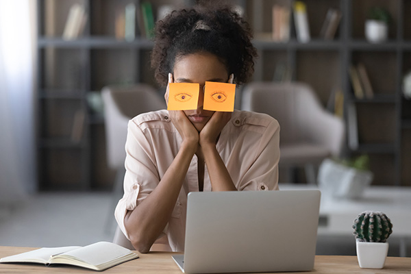 bored woman working on laptop with eyes covered in sticky notes with eyes drawn on them